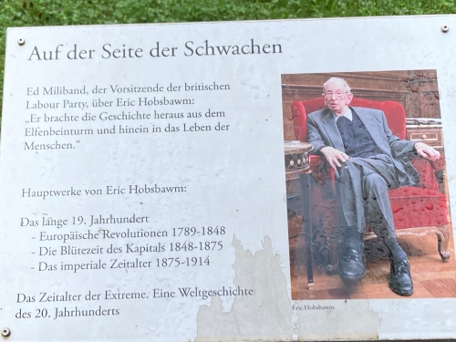 A photo of an information board, entitled ‘Auf der Seite der Schwachen’ [On the side of the weak], showing a colour photo of Eric Hobsbawm in his old age, with a quote from ‘Ed Miliband, leader of the British Labour Party’ about Hobsbawm leading History out of the Ivory tower and into people’s lives, and a short list of Hobsbawm’s major books.